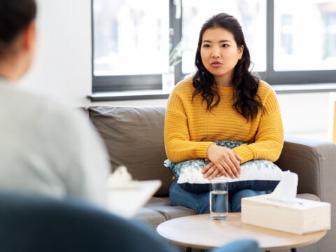 A young lady in a counselling session. She has dark hair and wears a yellow jumper. We only see the counsellor from behind, but they appear to be a woman with dark hair, wearing a white top.