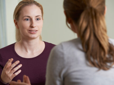 Two women talk. We only see one of their faces, she is blonde and is using her hands to express herself. We see the back of the head of the second woman, she has long brown hair, tied up in a ponytail.