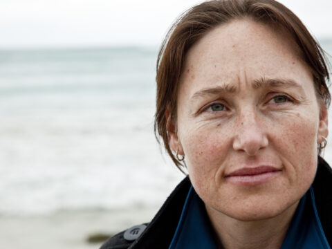 This image shows a close up of a woman's face, she is standing on a beach, with the shoreline behind her. She looks thoughtful.