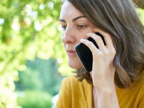 A woman on a phone call. The woman has shoulder length light brown hair and uses a modern mobile phone. She is wearing a yellow top. In the background are trees.